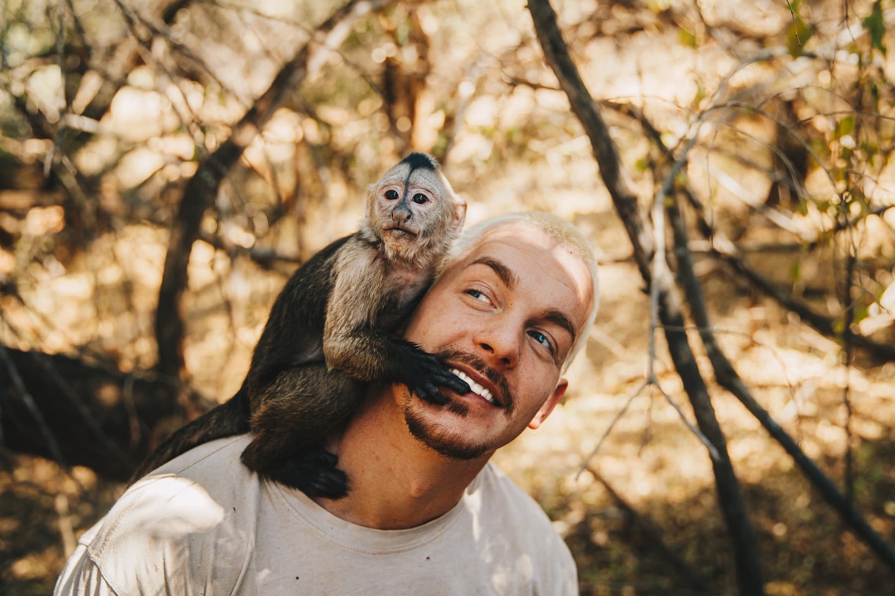 Dean is in the forest, wearing a white shirt. On his right shoulder sits a monkey. The monkey's name, which his sitting on his shoulder, is Momo and is one of the two monkey rescues on the Hakuna Mipaka Oasis.