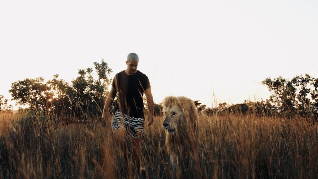 Dean is walking in the African wilderniss together his lion Snow. Snow is having a white fur, Dean is wearing a black shirt and short pants with a zebra pattern. 